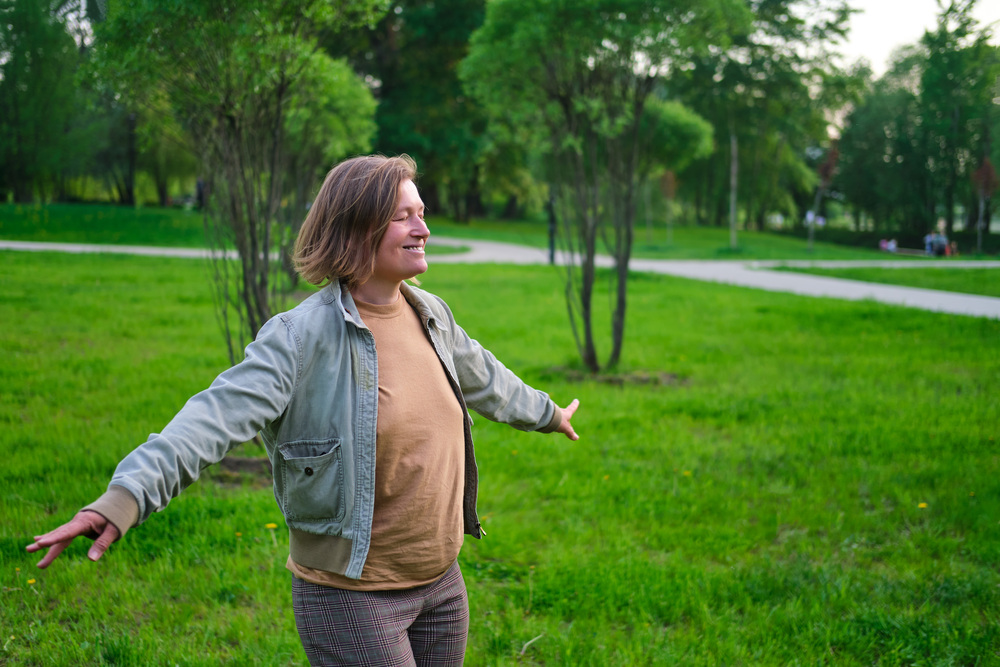 Person står med utsträckta armar i en grön park, ser glad ut och njuter av naturen.