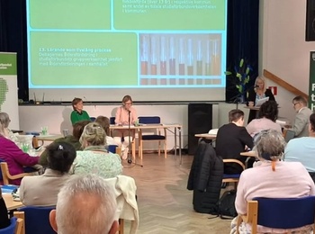 Konferens med deltagare som lyssnar på en presentation i en ljus sal med projektorduk och panel.