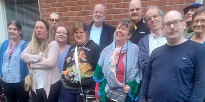 En grupp människor står och sitter utomhus framför en tegelvägg, några med färgglada kläder.