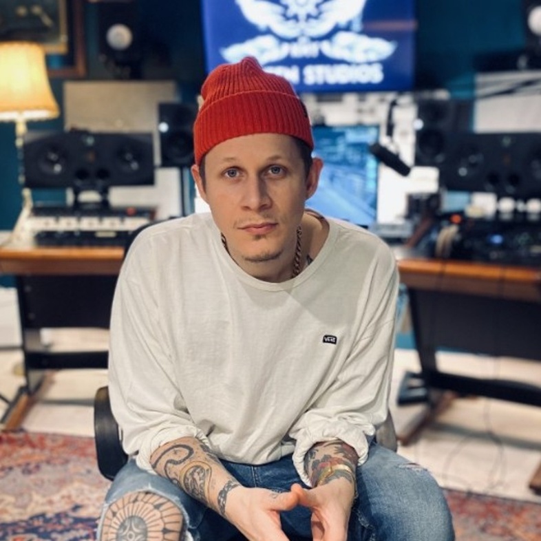 Man med röd mössa sitter i musikstudio med instrument och ljudutrustning runt sig.