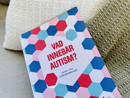 En bok med titeln 'Vad innebär autism?' ligger på en soffa med två kuddar bakom.