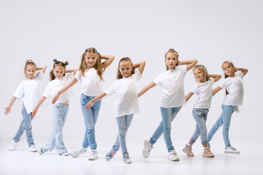 Sju unga flickor i vita t-shirts och jeans poserar i rad mot vit bakgrund.