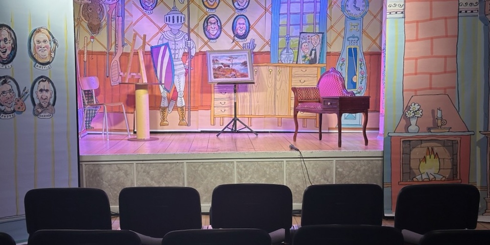 En liten teaterscen med färgglad kuliss som föreställer ett rum med riddarrustning, klocka och öppen spis. Stolar står framför scenen.