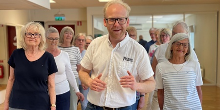 En grupp äldre personer och en yngre man står tillsammans inomhus, leende och avslappnade.