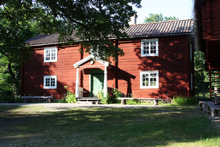 Röd trähus med vita fönster och grön dörr, omgiven av träd och gräsmatta i solsken.