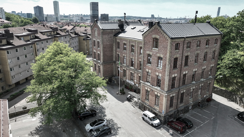 Tegelbyggnad med flera våningar, omgiven av parkerade bilar och träd, i ett bostadsområde med höghus i bakgrunden.