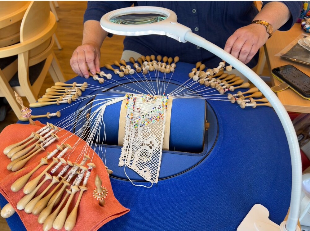 En person tillverkar spets med hjälp av knyppelpinnar, tråd och en förstoringslampa på ett blått bord.