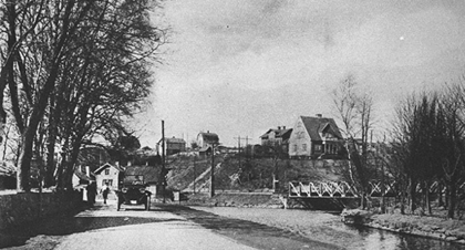 Gammalt svartvitt foto av en landsväg med träd, bro, hus och några människor och en bil.