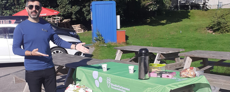 Man står bredvid ett picknickbord utomhus med grön duk, kaffe och fika under en solig dag.