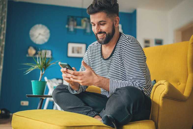 En person sitter i en gul fåtölj i ett vardagsrum och tittar på en mobil.