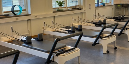 Ett ljust pilatesrum med flera reformermaskiner, stora fönster och träningsredskap på fönsterbrädorna.