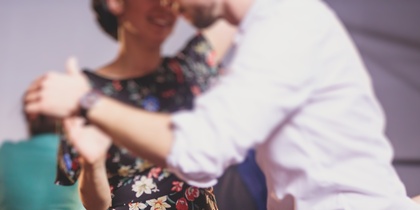 Två personer dansar tillsammans, en i blommig klänning och en i vit skjorta, i en suddig inomhusmiljö.