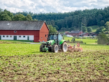En grön traktor plöjer en åker framför en röd ladugård på landsbygden med skog i bakgrunden.