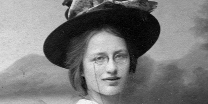 Svartvit foto av kvinna i långklänning och stor hatt, poserande framför målad bakgrund.