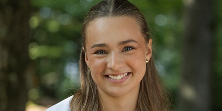 Ung kvinna med långt, vågigt hår och vit blus sitter utomhus och ler mot kameran.