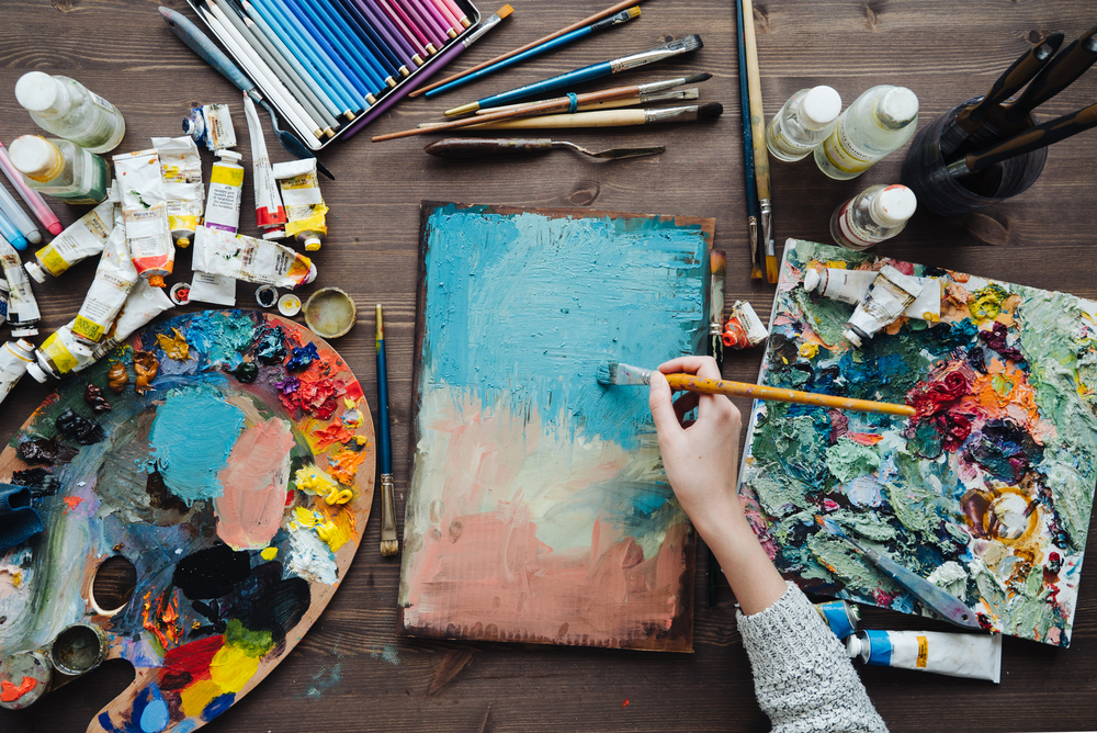 En person målar abstrakt konst på en duk med pensel, omgiven av färgtuber, penslar och färgpaletter.