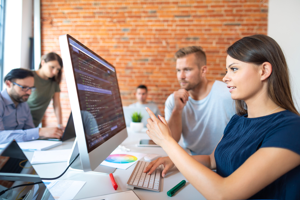 Fyra personer arbetar tillsammans vid datorer i ett modernt kontorslandskap med tegelvägg.