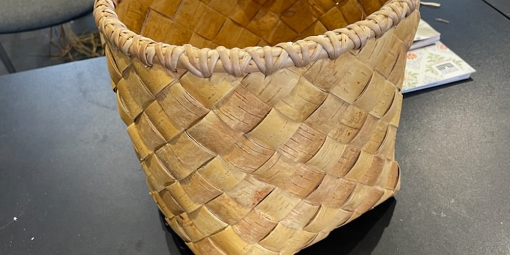 En handflätad korg av naturmaterial står på ett svart bord i ett rum med stolar och hantverksmaterial.