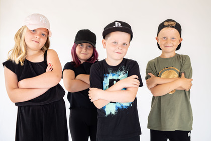 Fyra barn står i rad med armarna i kors och bär kepsar, mot en ljus bakgrund.