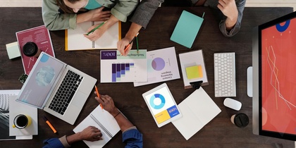 Tre personer arbetar tillsammans vid ett skrivbord med datorer, dokument och diagram.