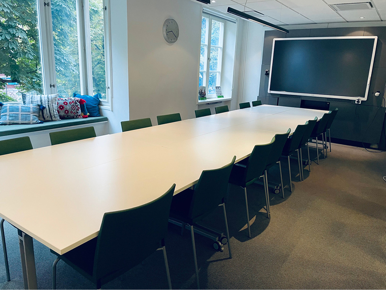 Möblerad lokal med bord och stolar. Stora fönster mot park.