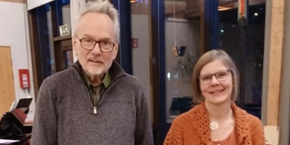 Två personer står inomhus bredvid ett piano, med stora fönster i bakgrunden.