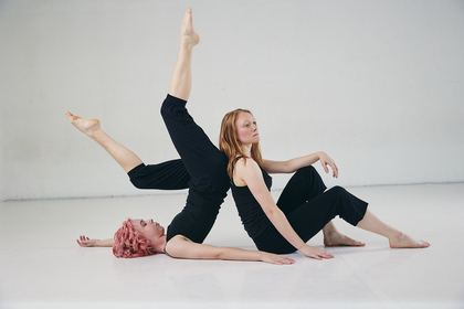 Två personer utför en modern danspose på golvet i ett ljust rum, klädda i svarta kläder.