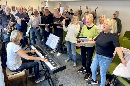 En grupp vuxna sjunger tillsammans i ett rum, en person spelar piano och en annan spelar saxofon.