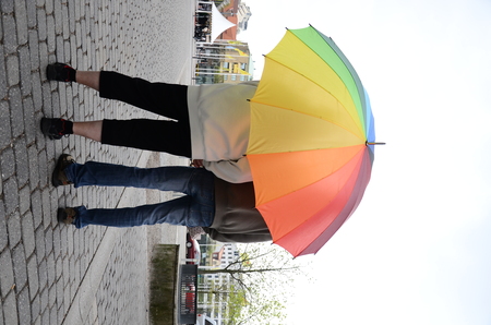 Två personer står under ett stort regnbågsfärgat paraply på en stenlagd gata, sedda bakifrån.