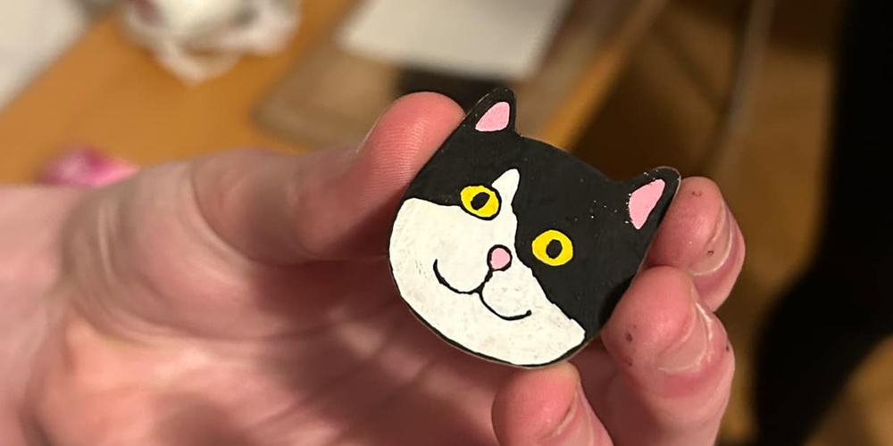 En hand håller en liten figur av en svartvit katt med gula ögon, i ett rum med trägolv och strykjärn i bakgrunden.