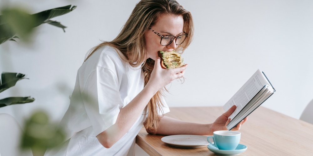 Kvinna med glasögon äter en smörgås och läser en bok vid ett träbord med en kopp kaffe.