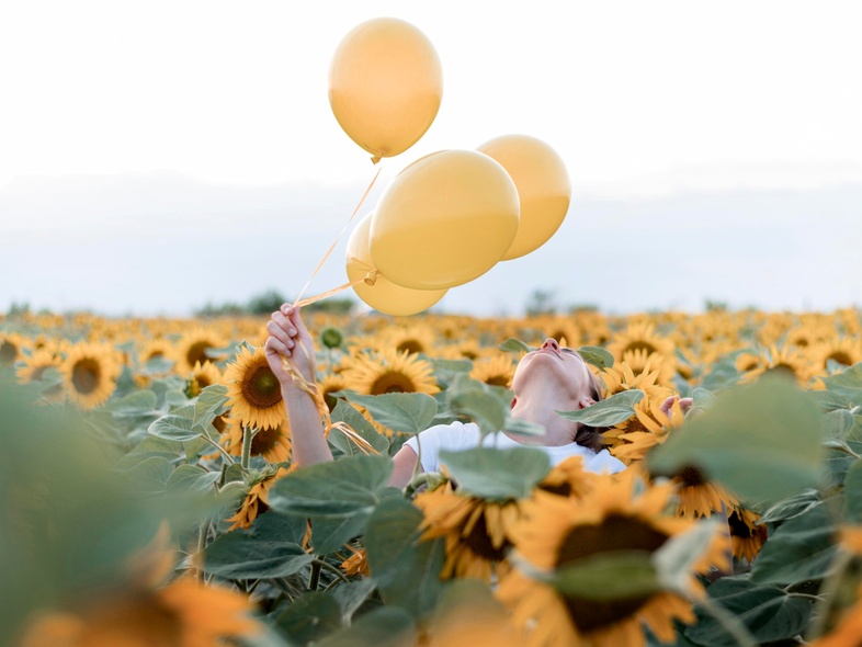 En person håller tre gula ballonger i ett fält av solrosor under en ljus himmel.