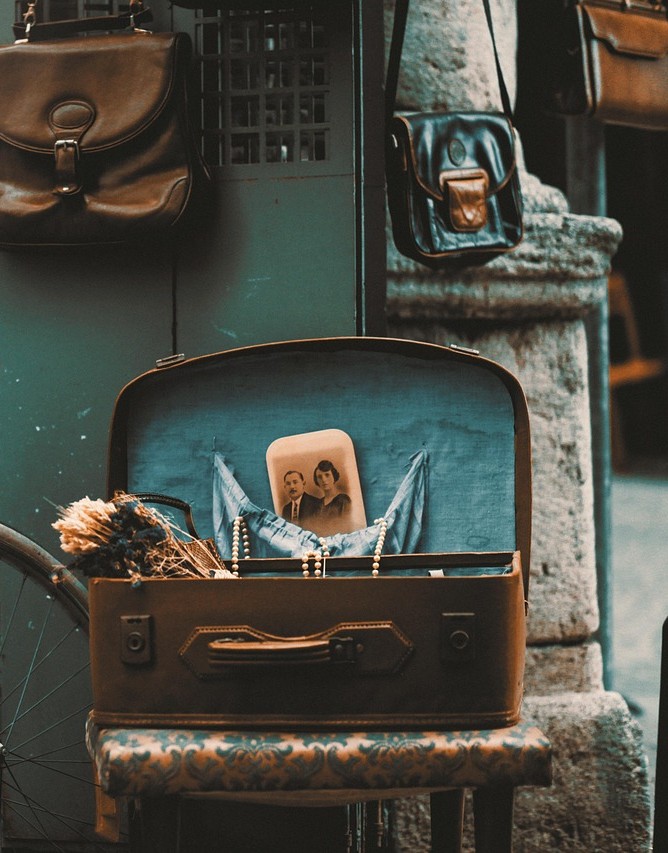En öppen vintage-resväska med ett gammalt svartvitt foto och torkade blommor, omgiven av väskor på en gata.