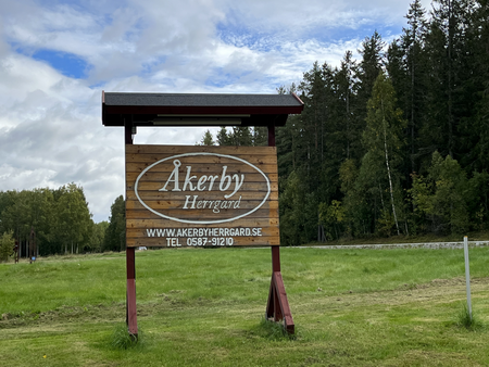Träskylt för Åkerby Herrgård står på en gräsmatta med skog i bakgrunden under en molnig himmel.