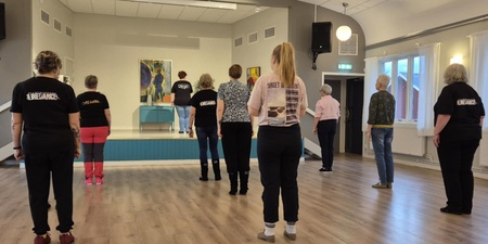 En grupp personer står i ett dansrum och följer en instruktör på en liten scen längst fram.