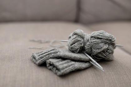 Grått garnnystan och stickade plagg med stickor ligger på en beige soffa.
