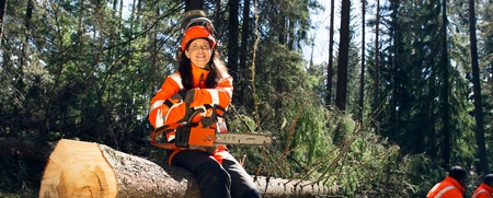 En person i orange skyddskläder sitter på en fälld trädstam i en skog med en motorsåg i händerna.