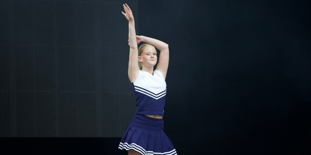 En ung kvinna i blå och vit cheerleaderdräkt står på scen med ena armen uppsträckt.