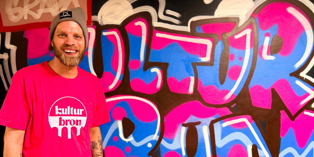 Person i rosa t-shirt med texten 'kultur bron' står framför färgglad graffiti-vägg.