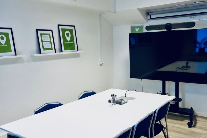 Ett modernt mötesrum med ett vitt bord, sex stolar, en stor skärm och tre tavlor på väggen.