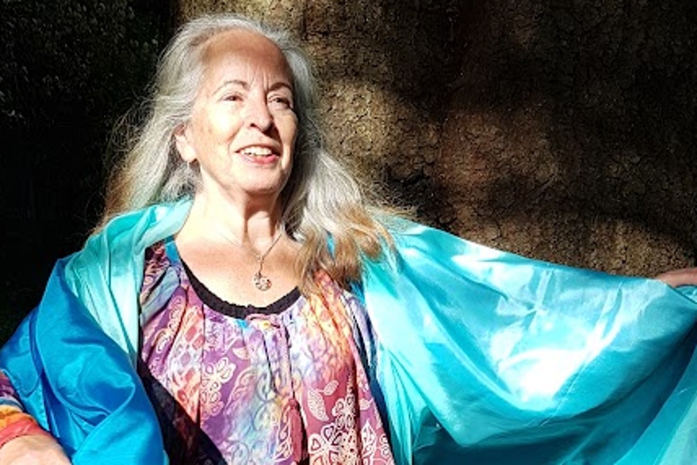 Cirkelledare Elisabeth Hjulström med långt grått hår står barfota utomhus, bär färgglad tunika och blå sjal.