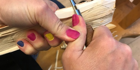 Närbild på händer som täljer trä med kniv. Naglarna är målade i olika färger.