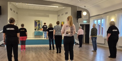 En grupp personer står i ett dansrum och följer en instruktör på en liten scen längst fram.