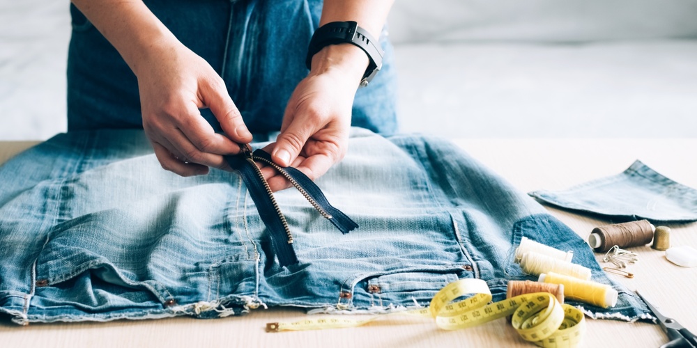 Person arbetar med att sy om ett par jeans, med dragkedja, måttband och sytråd på bordet.