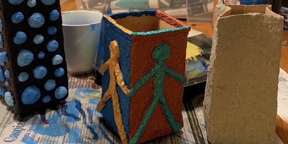 Tre färgglada, handgjorda pappersfigurer står på ett bord med konstmaterial och färg i bakgrunden.