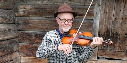 Äldre man med glasögon och hatt spelar fiol framför en vägg av trästockar.