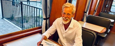 En äldre man med vitt hår och skägg sitter vid ett bord på ett tåg och håller en bok.