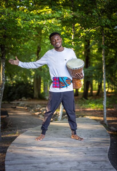 En leende man står barfota på en trästig i skogen och håller en trumma under armen.