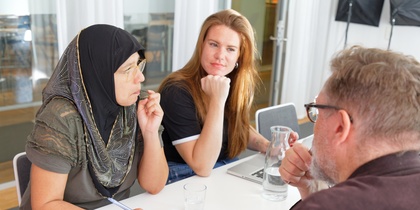 Tre personer sitter vid ett bord och diskuterar, med anteckningsblock och glas vatten framför sig.