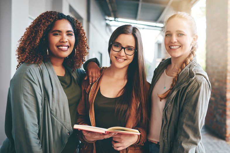 Tre unga kvinnor står tillsammans utomhus, leende och håller en bok.